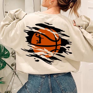 May include: A cream-colored sweatshirt with a graphic of a basketball with a player in silhouette. The basketball is orange and black and appears to be bursting through the fabric of the sweatshirt.