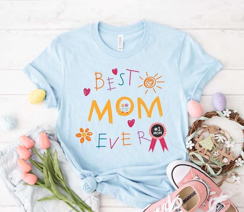May include: Light blue t-shirt with the text "Best Mom Ever" in colorful letters. The shirt also features a sun, flowers, hearts, and a red ribbon with the text "#1 Mom".