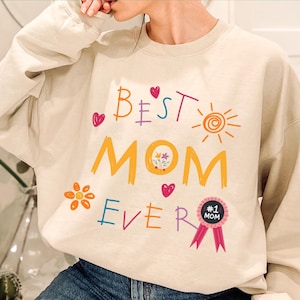 May include: A beige sweatshirt with the text "Best Mom Ever" in colorful, childlike handwriting. The text is surrounded by hearts, a sun, a flower, and a ribbon with the text "#1 Mom".