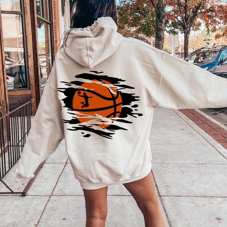 May include: A white hoodie with a basketball graphic on the back. The graphic shows a basketball with an orange and black design and a player in silhouette dunking the ball. The graphic is surrounded by black jagged lines, giving the impression of a ripped or torn fabric.