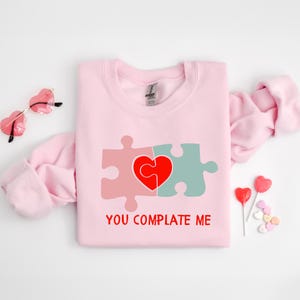 以下が含まれることがあります： ハートを形作る2つのパズルピースのグラフィックがプリントされたピンクのトレーナー。グラフィックの下には「You Complete Me」というテキストがプリントされています。
