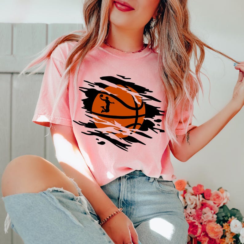 May include: A pink t-shirt with a graphic of a basketball with a player shooting a hoop. The basketball is in a ripped hole in the shirt.