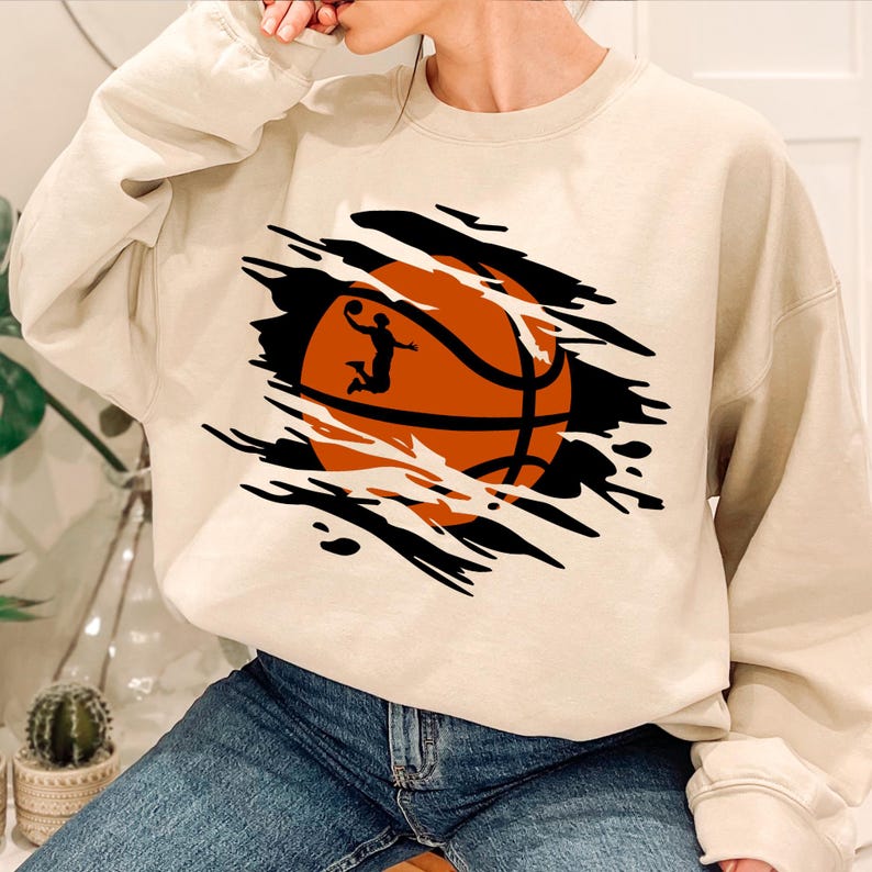 May include: A beige sweatshirt with a graphic of a basketball being ripped through the fabric. The basketball is orange and black with a silhouette of a basketball player dunking.