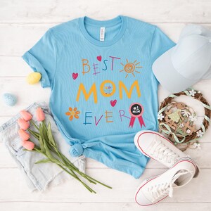 May include: A light blue t-shirt with the text "Best Mom Ever" in colorful, childlike handwriting. The shirt features a sun, flowers, and hearts. A pink ribbon with a black circle and the text "#1 Mom" is on the shirt.