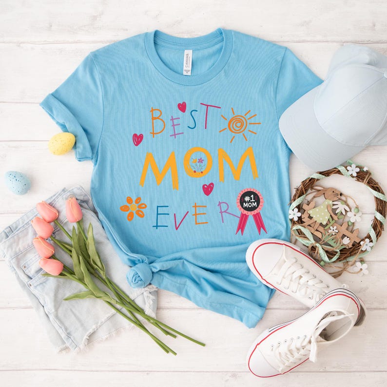 May include: A light blue t-shirt with the text "Best Mom Ever" in colorful, childlike handwriting. The shirt features a sun, flowers, and hearts. A pink ribbon with a black circle and the text "#1 Mom" is on the shirt.