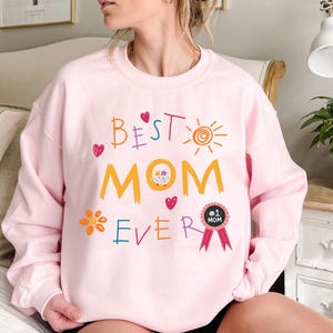 May include: Pink sweatshirt with the text "Best Mom Ever" in colorful, childlike handwriting. A flower, a heart, a sun, and a ribbon with the text "#1 Mom" are also included in the design.