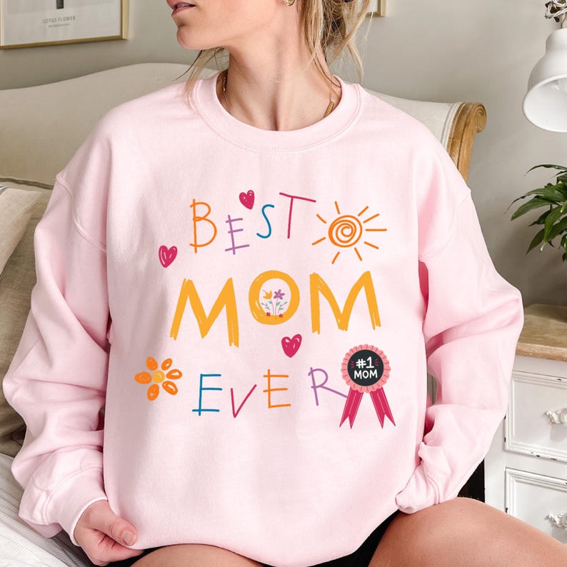 May include: Pink sweatshirt with the text "Best Mom Ever" in colorful, childlike handwriting. A flower, a heart, a sun, and a ribbon with the text "#1 Mom" are also included in the design.