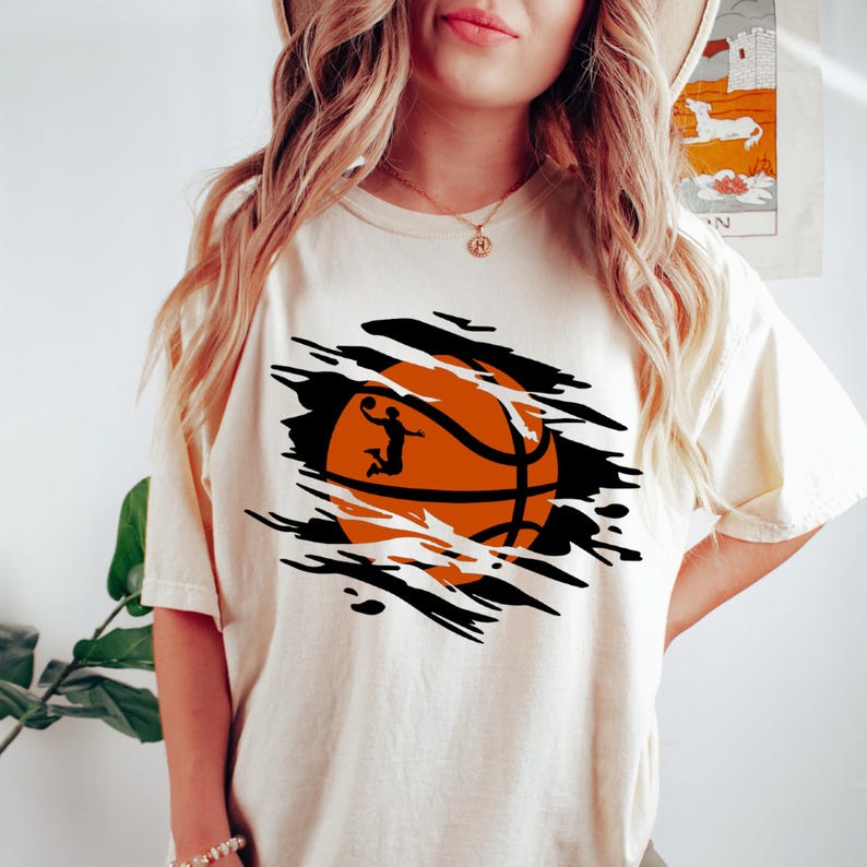 May include: A white t-shirt with a graphic of a basketball with a player dunking. The basketball is orange and black and is shown as if it is bursting through the shirt.