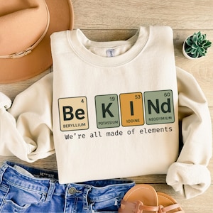 May include: A beige sweatshirt with a graphic print of the word "KIND" spelled out using the chemical symbols for Beryllium, Potassium, Iodine, and Neodymium. The text "We're all made of elements" is printed below the graphic.