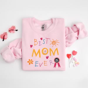 May include: Pink sweatshirt with the text "Best Mom Ever" in colorful, childlike handwriting. The text is surrounded by hearts, a sun, and a flower. A black and white ribbon with the text "#1 Mom" is below the text.