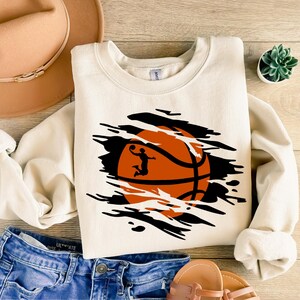 May include: A white sweatshirt with a black and orange basketball graphic. The basketball is ripped open to reveal a silhouette of a basketball player dunking.