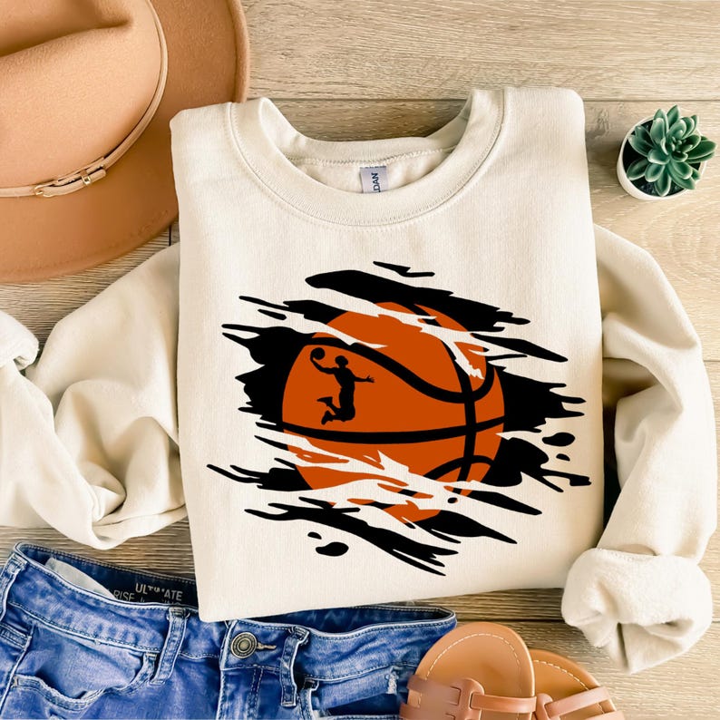 May include: A white sweatshirt with a black and orange basketball graphic. The basketball is ripped open to reveal a silhouette of a basketball player dunking.