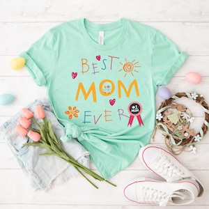 May include: A light green t-shirt with the text "Best Mom Ever" in colorful, childlike handwriting. The shirt features a sun, flowers, and hearts. A small black and white badge reads "#1 Mom".