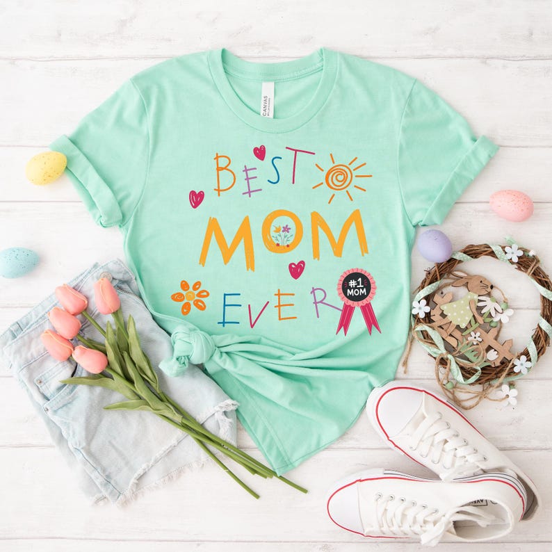 May include: A light green t-shirt with the text "Best Mom Ever" in colorful, childlike handwriting. The shirt features a sun, flowers, and hearts. A small black and white badge reads "#1 Mom".