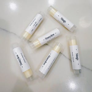 May include: Five lip balm tubes with white labels. The labels have black text that reads "Honeydew" and "Mango".