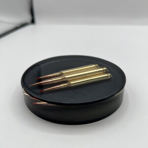 May include: Three gold-colored bullet casings with red tips are displayed on a black circular surface.
