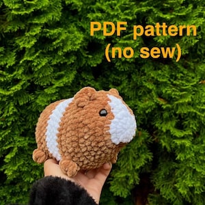 Pode incluir: Um brinquedo de pelúcia de cobaia de crochê marrom e branco. O brinquedo está sendo segurado na mão de uma pessoa. O texto "PDF pattern (no sew)" é exibido acima do brinquedo.