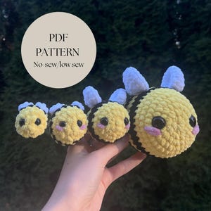 May include: Four crocheted bee plushies in different sizes, with yellow and black stripes, white wings, and black eyes. The plushies are arranged in a line, with the largest bee on the right and the smallest bee on the left. The plushies are held in a hand. The text "PDF PATTERN No-sew/low sew" is visible in the background.