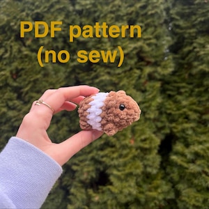 May include: A crocheted guinea pig in shades of brown and white, held in a hand. The text "PDF pattern (no sew)" is displayed in yellow above the guinea pig. The background is a blurred green.