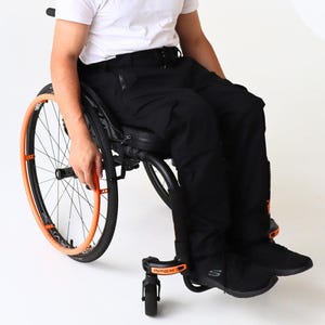 May include: A person in a wheelchair wearing a white V-neck shirt and black pants. The wheelchair has black and orange accents on the wheels and frame. The person is smiling.