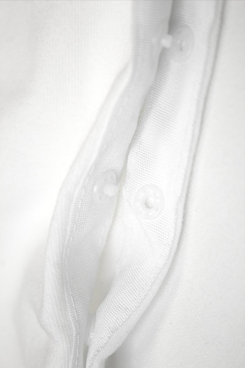 K&ouml;nnte beinhalten: Nahaufnahme eines wei&szlig;en Stoffes mit zwei transparenten Druckkn&ouml;pfen. Der Stoff scheint ein gewebtes Material zu sein, m&ouml;glicherweise Baumwolle oder Leinen, mit sichtbarer Textur. Die Druckkn&ouml;pfe sind gleichm&auml;&szlig;ig verteilt.