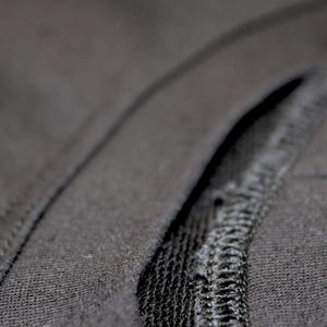 May include: Close-up of dark gray fabric with a visible seam. The fabric appears to be a soft knit material. The seam is stitched with a contrasting thread, creating a textured detail.