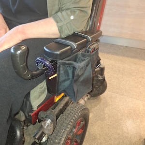 May include: A black wheelchair armrest with a padded armrest and two zipped pockets. The armrest is attached to a red wheelchair.