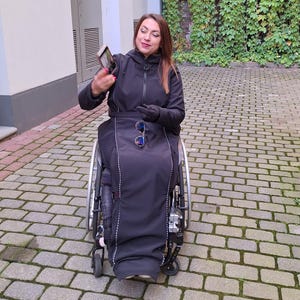 May include: A woman in a black jacket and black pants sits in a wheelchair. She is holding a smartphone in her right hand and looking at it. She is wearing sunglasses on her head.
