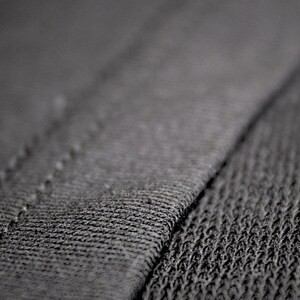 May include: Close-up of dark gray fabric with visible stitching. The texture of the material is rough and detailed, with a diagonal seam dividing the image. The fabric appears to be a knit or woven textile.