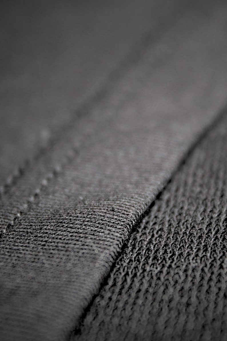 May include: Close-up of dark gray fabric with visible stitching. The texture of the material is rough and detailed, with a diagonal seam dividing the image. The fabric appears to be a knit or woven textile.