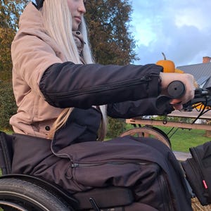 May include: A person in a wheelchair wearing a tan jacket with black sleeves and a black hat. The person is holding the handlebars of the wheelchair. A small orange pumpkin sits on a wooden cart in the background.