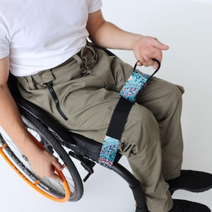 Può includere: Una sedia a rotelle nera con un cerchione arancione e una cinghia fantasia. La cinghia ha una base nera con un design colorato di forme blu, verde acqua e marrone. La persona indossa una camicia bianca e pantaloni color cachi.