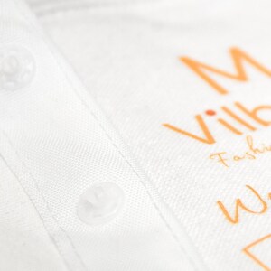 K&ouml;nnte beinhalten: Nahaufnahme eines wei&szlig;en Hemdes mit Kragen und sichtbaren Kn&ouml;pfen. Das Hemd hat einen orangefarbenen Text mit der Aufschrift "M Ville Fashion Wear". Der Stoff hat eine subtile Textur.