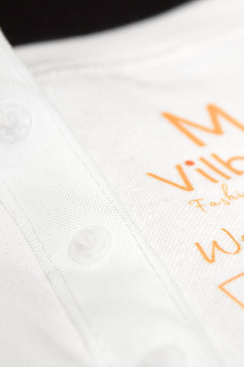 K&ouml;nnte beinhalten: Nahaufnahme eines wei&szlig;en Hemdes mit Kragen und sichtbaren Kn&ouml;pfen. Das Hemd hat einen orangefarbenen Text mit der Aufschrift "M Ville Fashion Wear". Der Stoff hat eine subtile Textur.