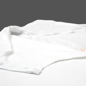 K&ouml;nnte beinhalten: Nahaufnahme eines wei&szlig;en Bodys mit Druckknopfverschluss. Das Kleidungsst&uuml;ck besteht aus einem weichen, wahrscheinlich Baumwollmaterial. Kragen und Schulterbereich sind sichtbar, mit einem Etikett in der oberen rechten Ecke.
