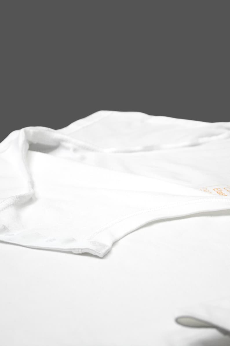 K&ouml;nnte beinhalten: Nahaufnahme eines wei&szlig;en Bodys mit Druckknopfverschluss. Das Kleidungsst&uuml;ck besteht aus einem weichen, wahrscheinlich Baumwollmaterial. Kragen und Schulterbereich sind sichtbar, mit einem Etikett in der oberen rechten Ecke.