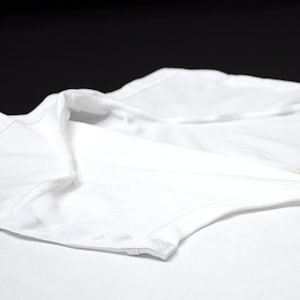 K&ouml;nnte beinhalten: Nahaufnahme eines wei&szlig;en Kleidungsst&uuml;cks, m&ouml;glicherweise ein Hemd oder Body, mit Kragen und Kn&ouml;pfen. Der Stoff scheint weich und leicht zu sein, mit sichtbaren N&auml;hten an den Kanten. Der Hintergrund ist einfarbig schwarz.