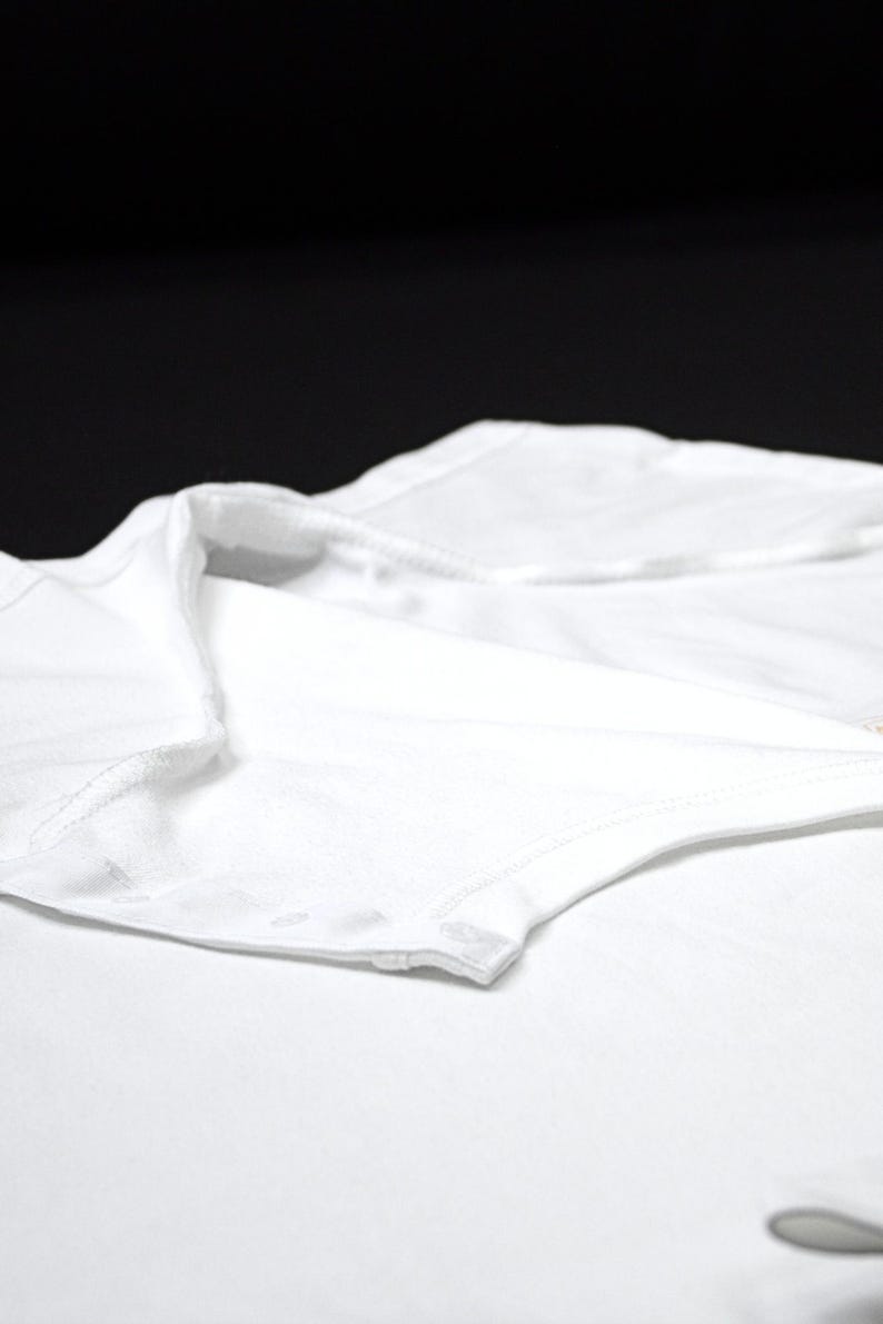 K&ouml;nnte beinhalten: Nahaufnahme eines wei&szlig;en Kleidungsst&uuml;cks, m&ouml;glicherweise ein Hemd oder Body, mit Kragen und Kn&ouml;pfen. Der Stoff scheint weich und leicht zu sein, mit sichtbaren N&auml;hten an den Kanten. Der Hintergrund ist einfarbig schwarz.