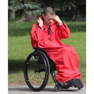Puede incluir: Un poncho rojo brillante para silla de ruedas con capucha y mangas largas. El poncho cubre a la persona en la silla de ruedas, extendiéndose hasta los reposapiés. La persona se está ajustando la capucha. El fondo es un parque con hierba verde y árboles.