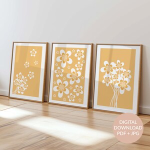 May include: Three framed prints with a yellow background and white stylised flower designs. The prints are arranged horizontally on a wooden floor.