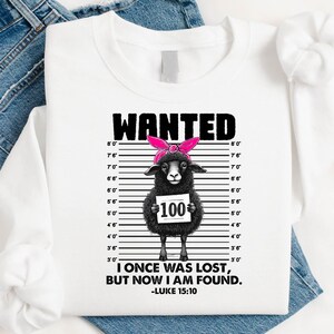 May include: White long-sleeve shirt with a graphic of a black sheep wearing a pink headband, holding a sign that says "100". The shirt has the word "WANTED" above the sheep and the text "I ONCE WAS LOST, BUT NOW I AM FOUND. -LUKE 15:10" below.