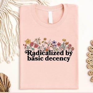May include: A heather peach-colored t-shirt with the text "Radicalized by basic decency" in black, above a floral design. The shirt is a crew neck style and is made of a soft material. The brand name "Elevated Ease" is at the bottom.