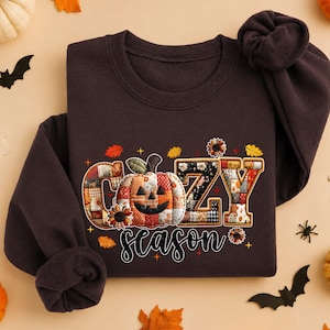 May include: A dark chocolate brown crewneck sweatshirt featuring a graphic print with the text "COZY season". The word "COZY" is filled with a patchwork pattern of autumn colors, and the letter "O" is replaced by a pumpkin illustration.