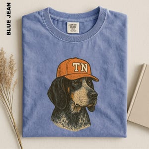 May include: A blue jean colored t-shirt featuring a graphic illustration of a dog wearing an orange baseball cap with the letters TN printed on the front. The shirt has a crew neckline and a Comfort Colors brand label on the inside collar area.