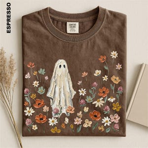May include: An espresso-colored short-sleeve t-shirt featuring a central graphic of a white ghost surrounded by a variety of colorful painted-style flowers in shades of orange, pink, yellow, and white. The shirt has a crew neckline and a label.