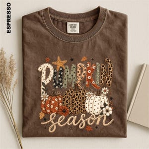 May include: An espresso-colored short-sleeve t-shirt featuring a graphic print with the text PUMPKIN season. The design includes three pumpkins with various patterns like leopard print, polka dots, and floral motifs, accented by a star and leaves.