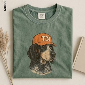 May include: A moss green crew neck t-shirt featuring a graphic illustration of a dog wearing an orange baseball cap with the letters TN printed on the front. The shirt has a visible brand label on the inside collar and a textured fabric finish.