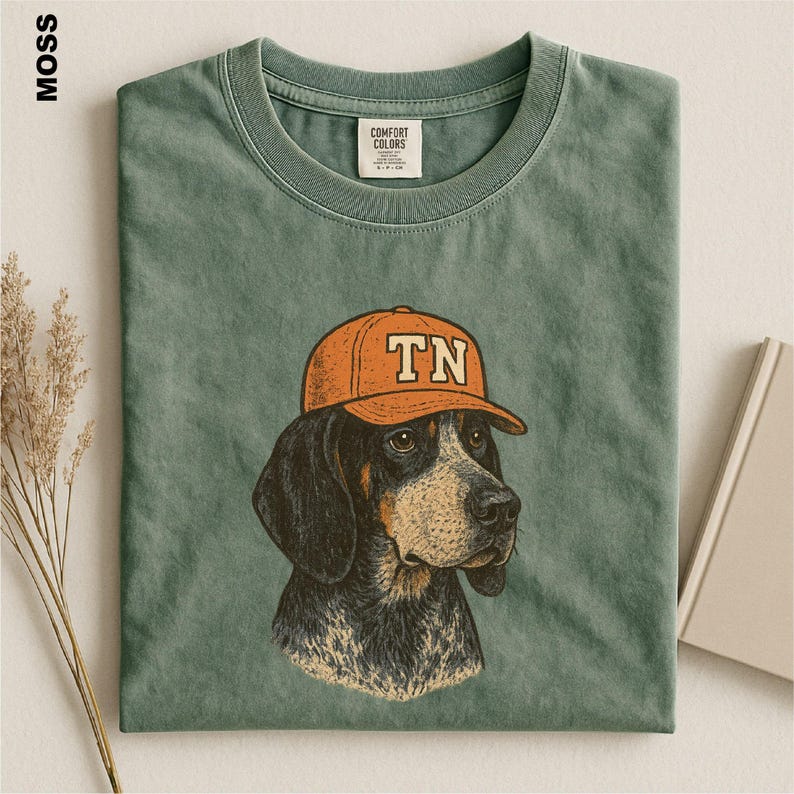 May include: A moss green crew neck t-shirt featuring a graphic illustration of a dog wearing an orange baseball cap with the letters TN printed on the front. The shirt has a visible brand label on the inside collar and a textured fabric finish.