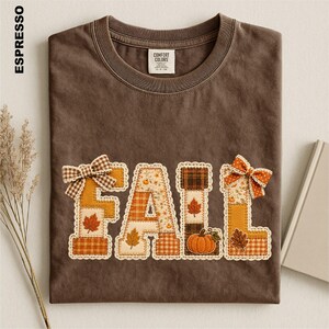 May include: A dark brown crew neck t-shirt featuring the word FALL in large, block-style letters. Each letter is filled with various autumn-themed patterns, including plaid, gingham, and leaf prints, accented with small fabric bows and a pumpkin.