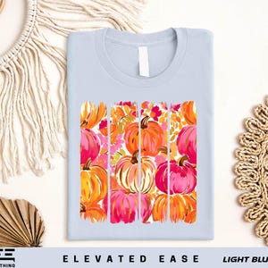 a light blue t-shirt with a vibrant graphic design of orange and pink pumpkins, arranged in a grid pattern. The design is displayed on a white background, and there are additional decorative elements such as a woven basket and a macrame wall hanging in the background.